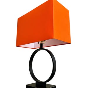 Pode incluir: Um candeeiro de mesa com um abajur retangular de tecido laranja e uma base de metal preto. A base consiste numa peça inferior retangular sólida que suporta uma estrutura circular ligada ao casquilho por baixo do abajur.