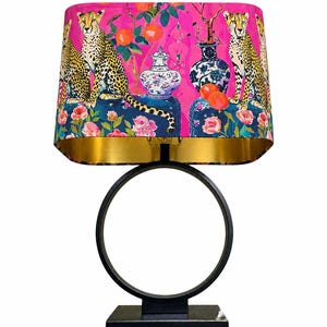 May include: A table lamp with a black circular base and stand. The drum lampshade features a vibrant pink background with a repeating pattern of cheetahs, blue and white porcelain vases, orange fruit, and pink flowers, finished with a gold interior.