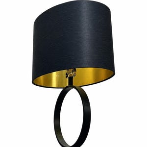 Op de afbeelding: Een tafellamp met een zwarte ovale voet en een bijpassende zwarte cilindrische lampenkap met een metallic gouden binnenvoering. De lamp heeft een centrale verticale ringstructuur die de voet verbindt met de fitting.