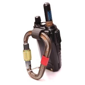 May include: A black two-way radio with a black antenna attached to the top. The radio is secured to a brown carabiner with a red strap.