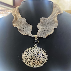 Peut inclure: Un collier en argent avec un grand pendentif rond orné d'une pierre noire et d'un motif de filigrane complexe. Le collier est fait d'une chaîne délicate et tissée.