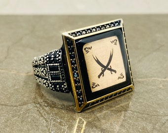 May include: A silver ring with a black and gold square design. The square features a gold background with two crossed swords in black. The ring is decorated with black stones.