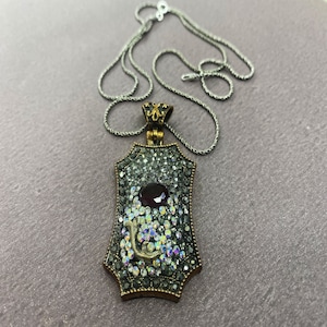 May include: A silver chain necklace with a pendant featuring a large, ornate, gold-toned frame encrusted with clear and black rhinestones. A single, dark red gemstone is set in the center of the frame.