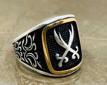 May include: A silver ring with a black enamel center featuring a silver design of two crossed swords. The ring has a gold trim around the enamel center.