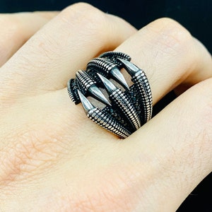 Peut inclure: Une bague en argent avec un motif de griffe. Les griffes sont pointues et ont des accents noirs. La bague est portée au doigt d'une personne.