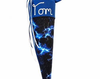May include: A cone-shaped school gift bag featuring a black fabric body with a blue lightning bolt pattern. The top section is solid blue fabric with the name Tom embroidered in white script, finished with white ribbon ties at the closure.