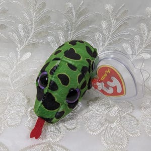 May include: A green plush snake toy with black spots, large purple eyes, and a red tongue. The toy has a clear heart-shaped tag attached. The tag reads "Teeny Tys" and features a red heart logo.