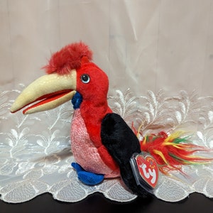 Peut inclure: Un oiseau en peluche rouge, noir et jaune avec un long bec et une étiquette Ty Beanie Baby. L'oiseau est assis sur une nappe en dentelle blanche.