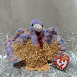 Peut inclure: Dinde en peluche à motif violet et orange avec une barbe rouge. La dinde est assise sur une surface blanche avec une étiquette Ty Beanie Babies attachée à son aile.