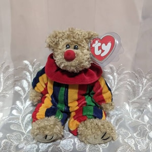 Peut inclure: Un ours en peluche clown brun et beige portant une combinaison rayée multicolore avec un col rouge. L'ours a un nez rouge et une étiquette Ty attachée à son oreille.