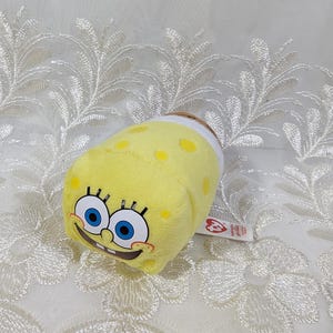 May include: Yellow plush toy of SpongeBob SquarePants wearing a white beanie. The toy is square shaped and has a smiling face with blue eyes. The toy has a Ty tag attached to it.
