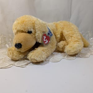 May include: A soft, plush golden retriever dog toy in shades of yellow and cream, with a black nose and a red heart-shaped tag. The toy is lying down. It has a blue tag attached to its ear.