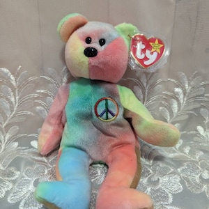 May include: A multi-colored tie-dye beanie baby bear with a peace sign embroidered on its chest. The bear has a red Ty tag with the words "Beanie Baby" and the Ty logo.