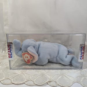 May include: A light blue plush elephant beanie baby toy in a clear plastic display case. The toy has a red heart-shaped tag with the number "3" on it. The case has "BEANS" printed on the side.