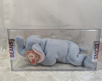 May include: A light blue plush elephant beanie baby toy in a clear plastic display case. The toy has a red heart-shaped tag with the number "3" on it. The case has "BEANS" printed on the side.