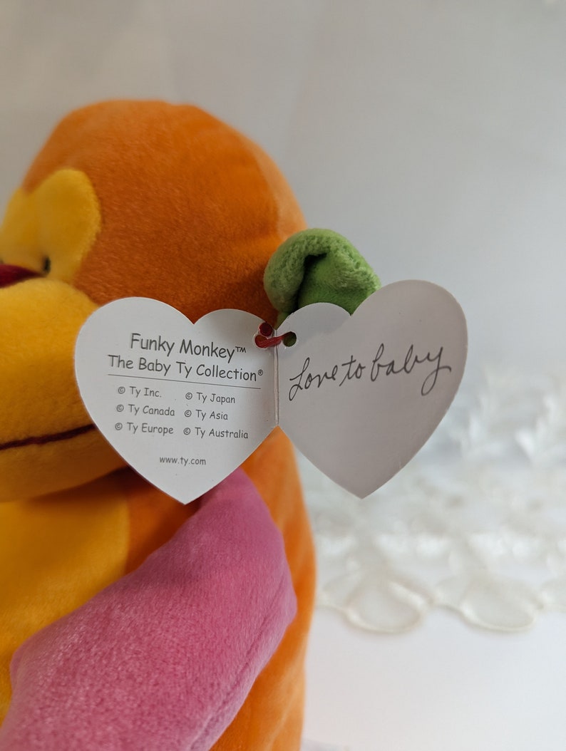 Pode incluir: Brinquedo de peluche de macaco laranja, amarelo e rosa com uma folha verde na cabe&ccedil;a. O macaco est&aacute; segurando uma etiqueta em forma de cora&ccedil;&atilde;o que diz "Love to baby" e tem o texto "Funky Monkey&trade; The Baby Ty Collection&reg; Ty Inc. &copy; Ty Japan Ty Canada &copy; Ty Asia Ty Europe &copy; Ty Australia www.ty.com".