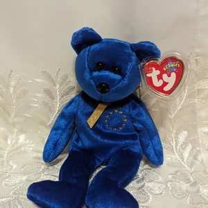 May include: A blue plush bear with a gold ribbon and a tag. The bear has a black nose and eyes. The tag is red with a heart shape and the word "Beanie Babies" printed on it.