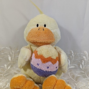 May include: A yellow plush duck holding a purple, orange, and white Easter egg. The duck has orange feet and a yellow beak.