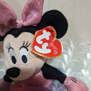 以下が含まれることがあります： ピンクのリボン、ドレス、手袋をつけたミニーマウスのぬいぐるみ。おもちゃは黒い耳と「Sparkle」と「Disney」と書かれた赤いハート型のタグが付いています。おもちゃは柔らかい、ぬいぐるみ素材で作られています。