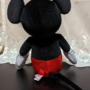 May include: A black plush Mickey Mouse toy with red shorts and a black tail. The toy is sitting on a black surface.