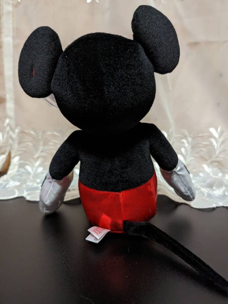 May include: A black plush Mickey Mouse toy with red shorts and a black tail. The toy is sitting on a black surface.