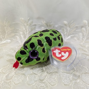 May include: A green plush snake toy with black spots, purple glitter eyes, and a red tongue. The toy has a "Teeny Tys" tag attached. The snake is lying on a white, patterned surface.