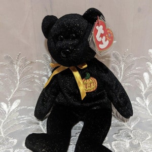 May include: Black glitter teddy bear plush toy with a yellow ribbon and a pumpkin patch. The bear has a red heart-shaped tag attached. A Halloween-themed collectible.