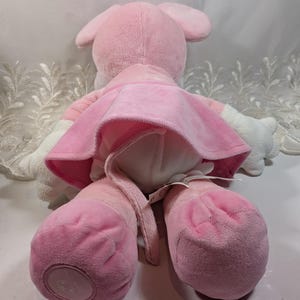 May include: Pink plush mouse wearing a pink dress. The mouse has a long tail and is sitting on a white surface.