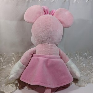 May include: Pink plush mouse toy with a pink skirt and white arms. The mouse has a long tail and is sitting on a white surface.
