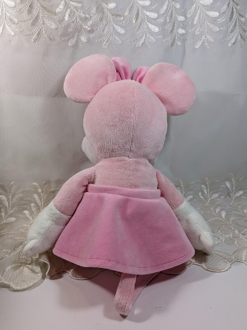 May include: Pink plush mouse toy with a pink skirt and white arms. The mouse has a long tail and is sitting on a white surface.