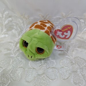 May include: A small, plush turtle toy with a green head and a brown and white spotted shell. The turtle has large, glittery eyes and a Ty heart-shaped tag attached. The toy is designed for children.