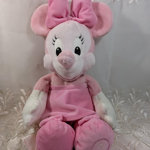 May include: Pink plush Minnie Mouse toy with a pink bow, white gloves, and a pink dress. The toy is sitting on a white lace surface.
