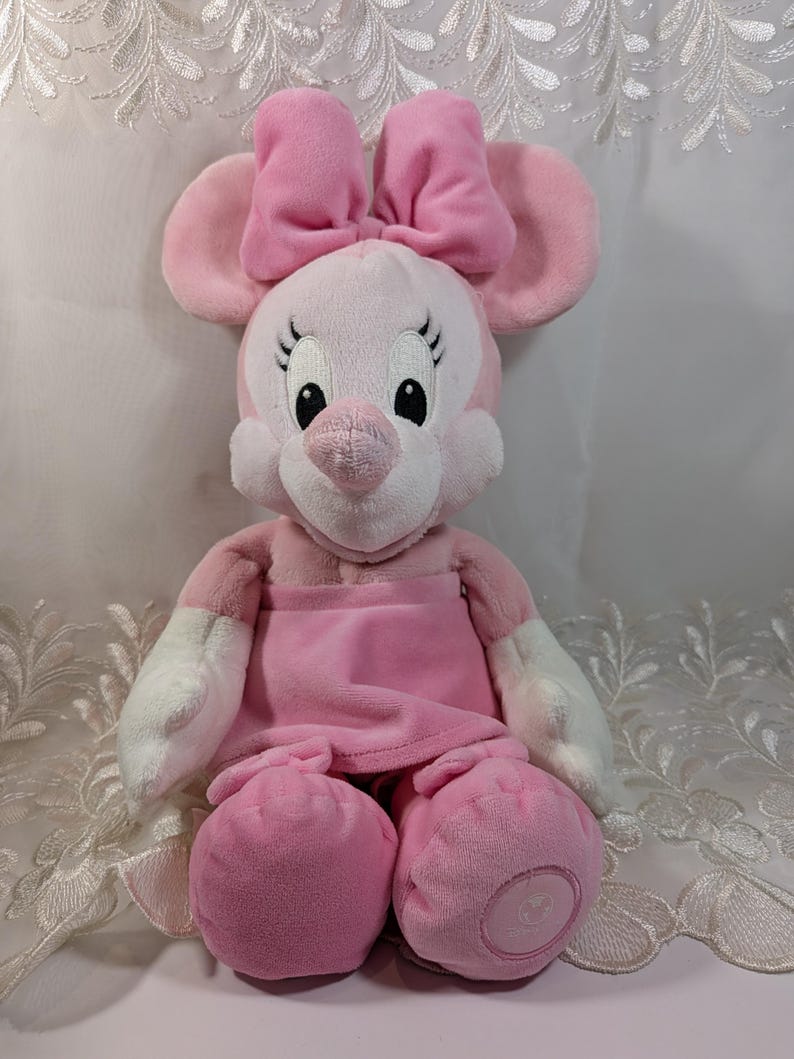 May include: Pink plush Minnie Mouse toy with a pink bow, white gloves, and a pink dress. The toy is sitting on a white lace surface.