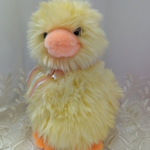 May include: A yellow fluffy plush duck with a pink beak and orange feet. The duck is wearing a pink and white ribbon around its neck.