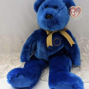May include: A blue teddy bear with a gold ribbon and a circular emblem with gold stars. The bear has a TY heart-shaped tag. The bear is sitting on a white surface.