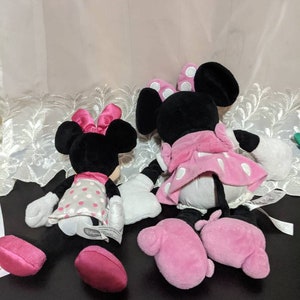 May include: Two plush Minnie Mouse toys, one in a pink and white polka dot dress and the other in a grey and white polka dot dress. Both toys have pink bows and pink shoes.
