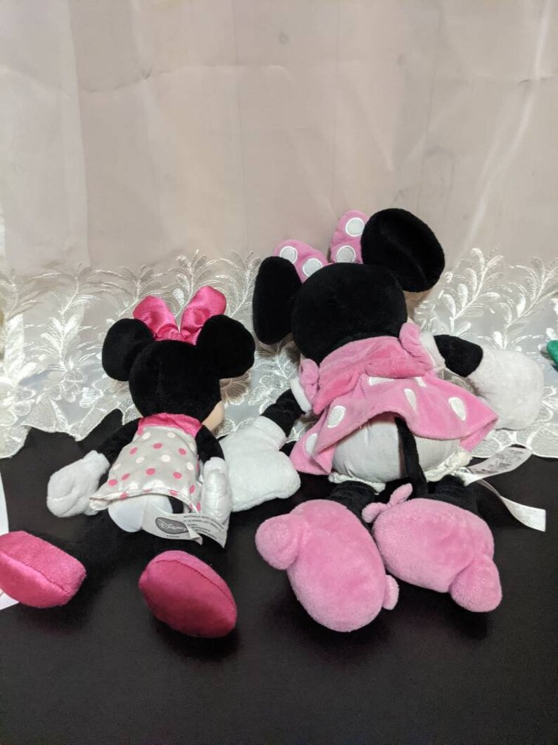 May include: Two plush Minnie Mouse toys, one in a pink and white polka dot dress and the other in a grey and white polka dot dress. Both toys have pink bows and pink shoes.