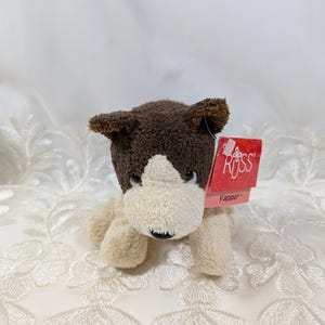 May include: A soft toy dog with brown and cream fur. The dog has a brown head and ears, a cream face and body, and black eyes. A red tag reads "RUSS Yapper".