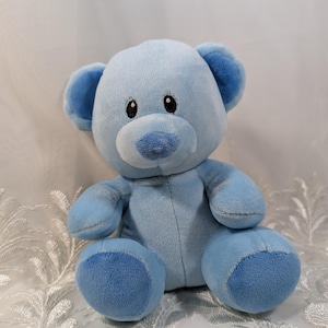 May include: A plush, light blue teddy bear with darker blue accents on the ears, nose, and paws. The bear has black button eyes and a soft, cuddly appearance. The toy is designed for children.
