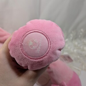 May include: Pink plush toy with a white embroidered Disney logo on the bottom. The toy has a bow on the back.