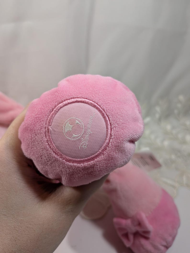 May include: Pink plush toy with a white embroidered Disney logo on the bottom. The toy has a bow on the back.