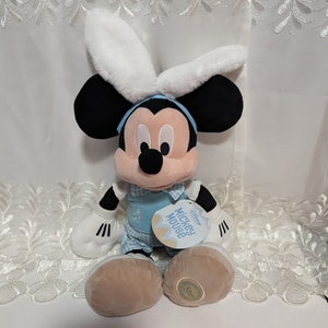 May include: A plush Mickey Mouse wearing a blue and white Easter outfit with bunny ears. The plush toy is sitting on a white lace background. The tag reads "Disney Mickey Mouse Easter Fashion".