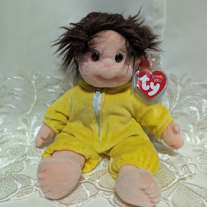 May include: A yellow plush toy with brown hair, wearing a yellow jumpsuit with a zipper. The toy has a pink face with brown eyes and a big smile. The toy has a Ty Beanie Kids tag attached to its arm.