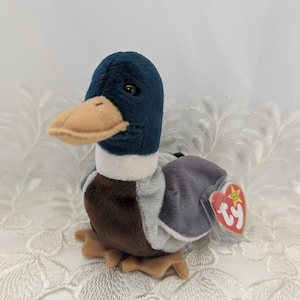 May include: A blue, brown and grey stuffed duck with a white stripe around its neck. The duck has a Ty tag attached to its wing.