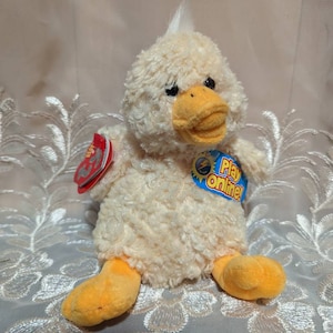 May include: A plush yellow duck with orange feet and a white tuft of hair on its head. The duck is sitting on a white lace tablecloth. The duck has a tag that says "Play Online!"