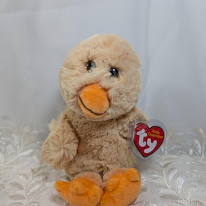 May include: A plush, light brown duck toy with an orange beak and feet. The duck has black eyes and a red heart-shaped tag that reads "Attic Treasures" by Ty.