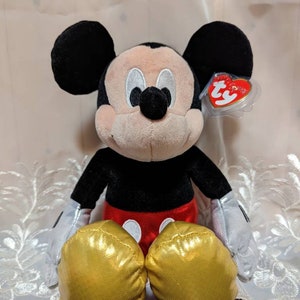 May include: A plush Mickey Mouse toy sitting with his legs crossed. He is wearing a red and black outfit with gold shoes. The toy has a Ty tag attached to his ear.