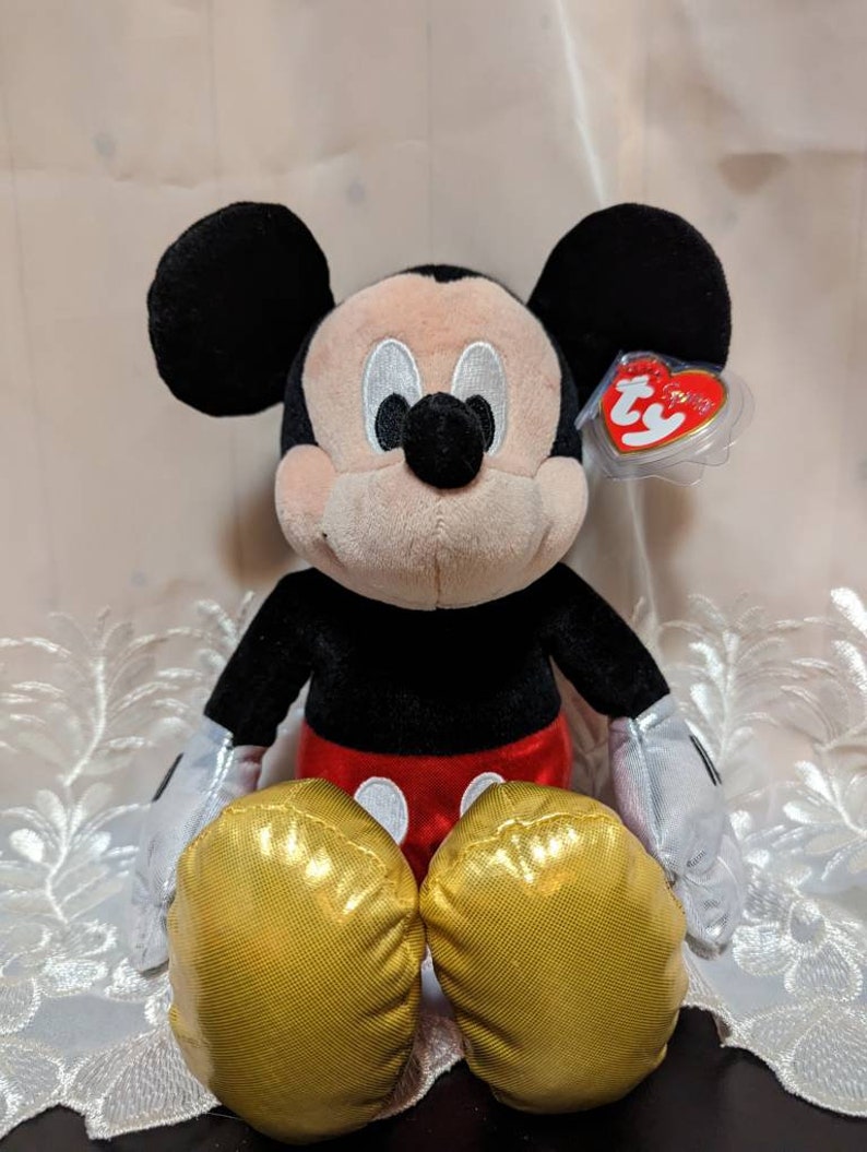 May include: A plush Mickey Mouse toy sitting with his legs crossed. He is wearing a red and black outfit with gold shoes. The toy has a Ty tag attached to his ear.