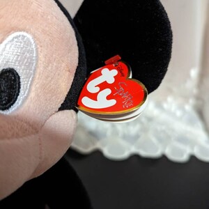 May include: A black and red Ty Beanie Baby plush toy of Mickey Mouse. The toy has a red heart-shaped tag with the words "Ty" and "Sparkles" on it.