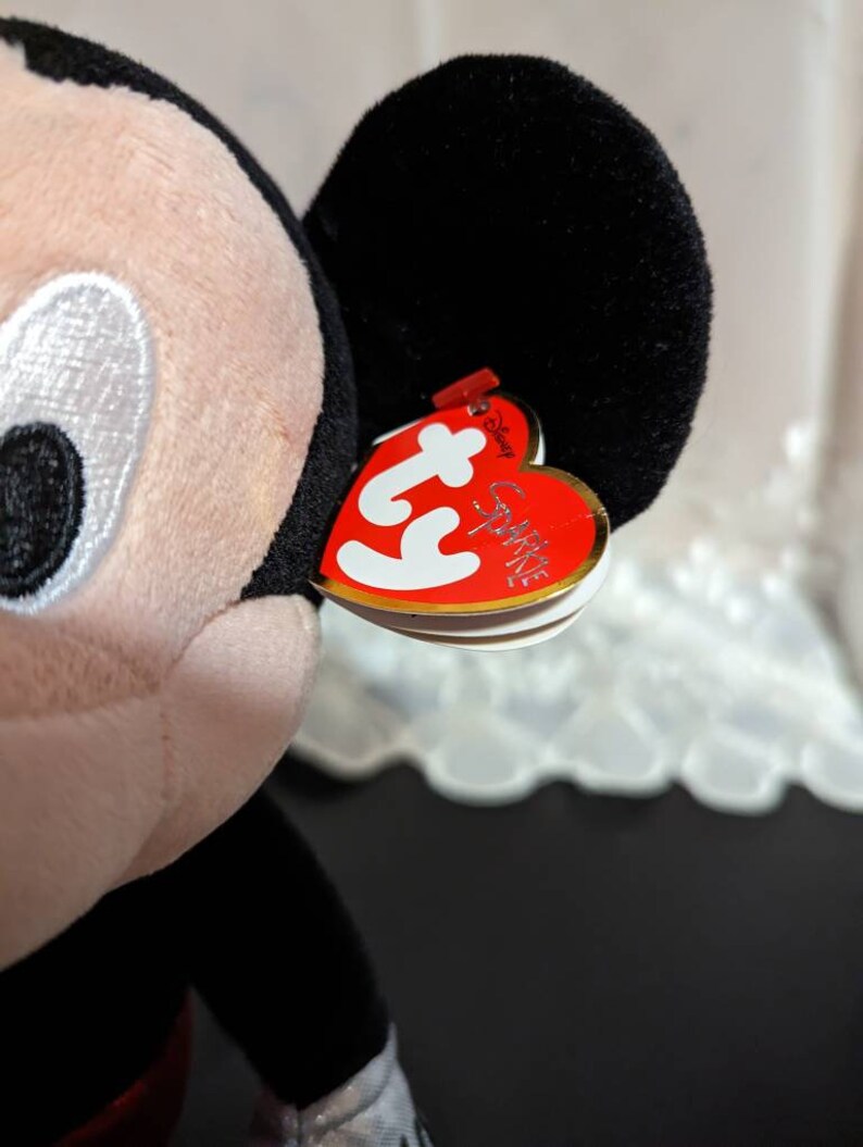 May include: A black and red Ty Beanie Baby plush toy of Mickey Mouse. The toy has a red heart-shaped tag with the words "Ty" and "Sparkles" on it.